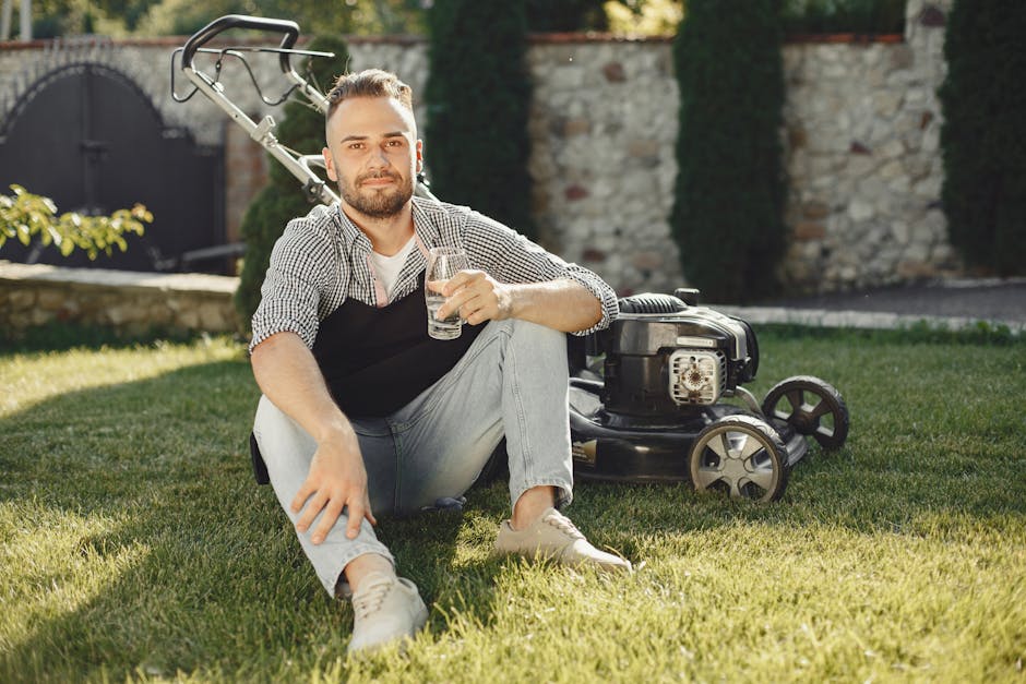 A person operating a mechanical lawn mower on a lush green lawn during a sunny day, with the camera positioned low to the ground capturing the freshly cut grass blades in the foreground. The mower's e